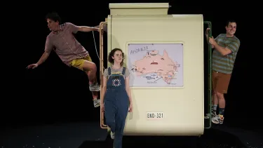 Two men standing on the ladder of a camper trailer with a woman in front of it.